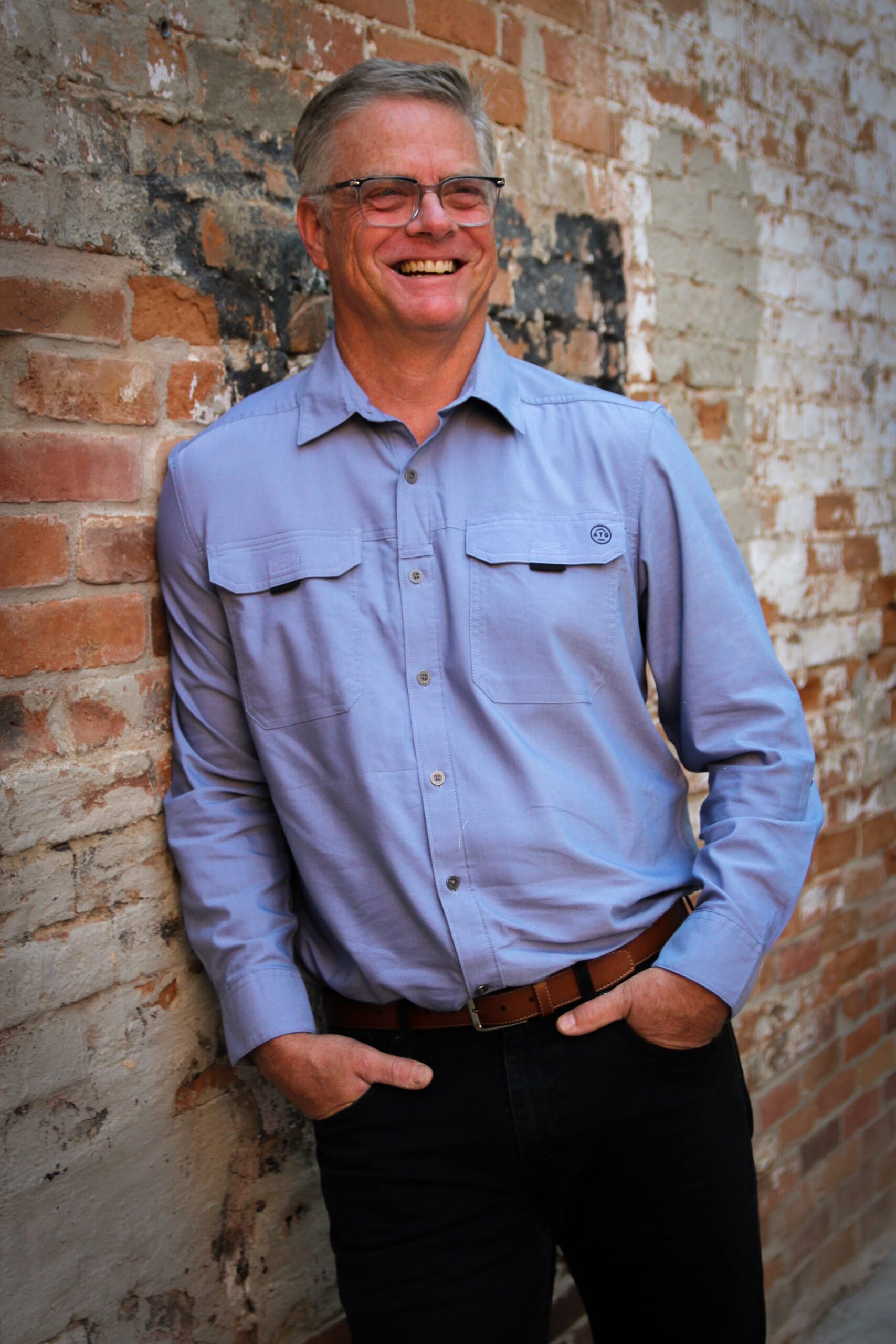 A photographic portrait of Kirk Romberg smiling and standing outside in front of a mottled red brick wall